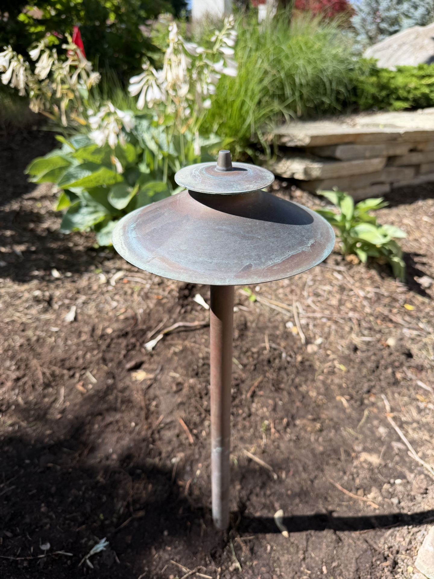 Small rusted garden path light in mulch, with stacked stone border and green plants behind it. Small rusted garden path light in mulch, with stacked stone border and green plants behind it.