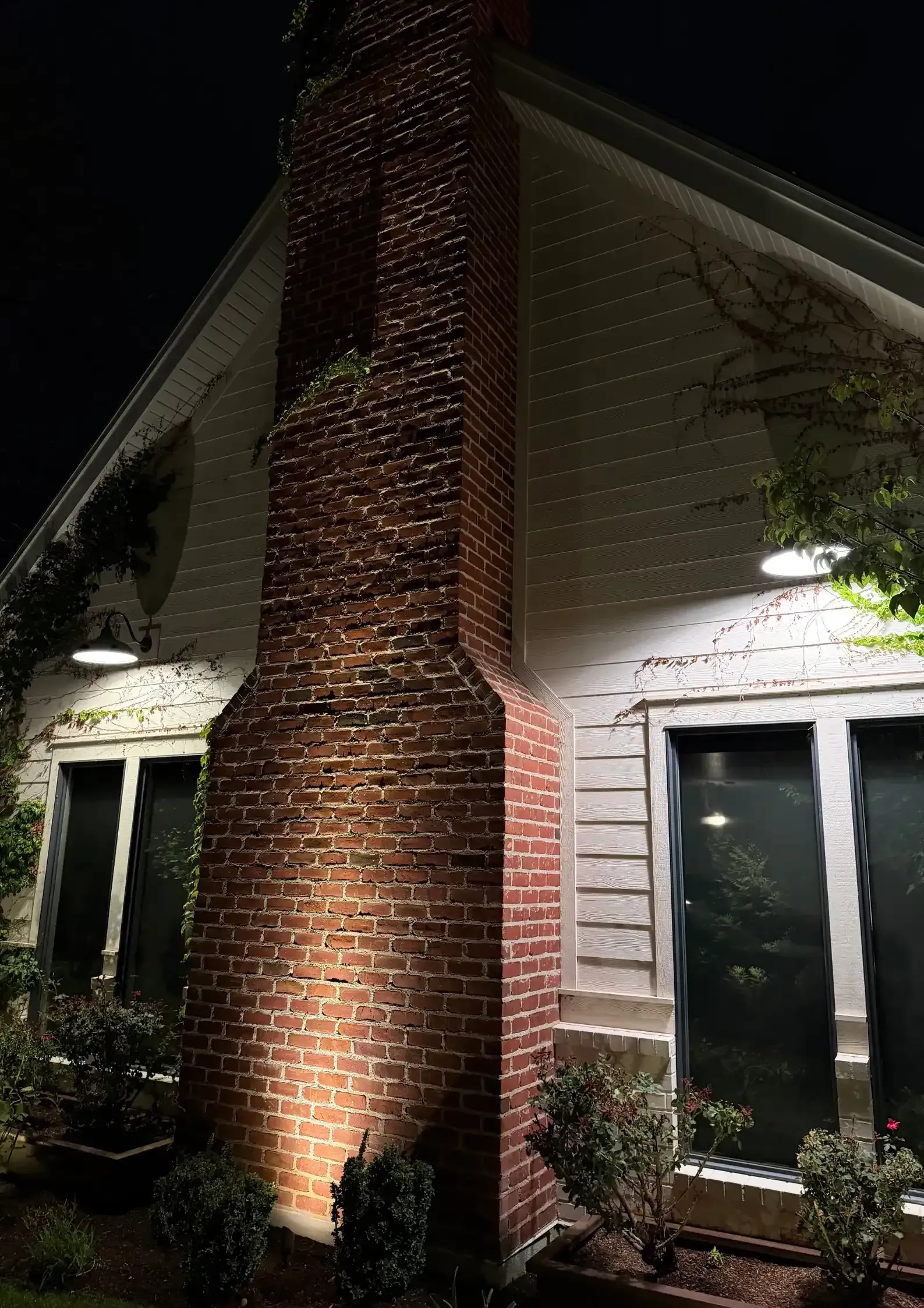 House exterior at night with a brick chimney lit by warm lights and two windows on either side House exterior at night with a brick chimney lit by warm lights and two windows on either side