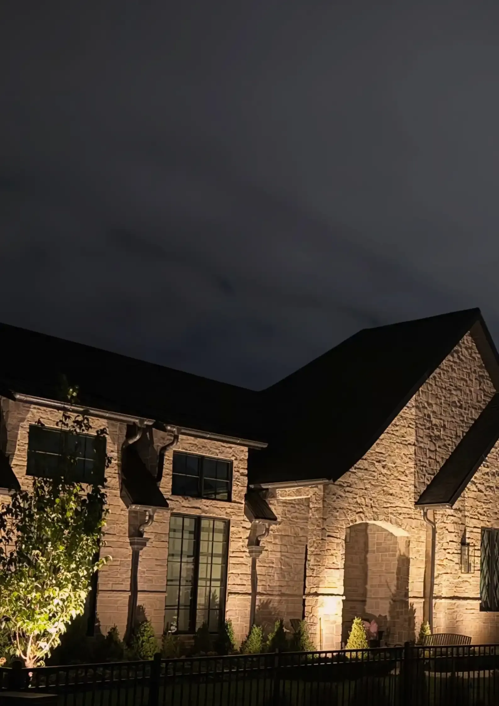Stone house lit at night under a cloudy sky, with a dark roof and glowing facade lights. Stone house lit at night under a cloudy sky, with a dark roof and glowing facade lights.