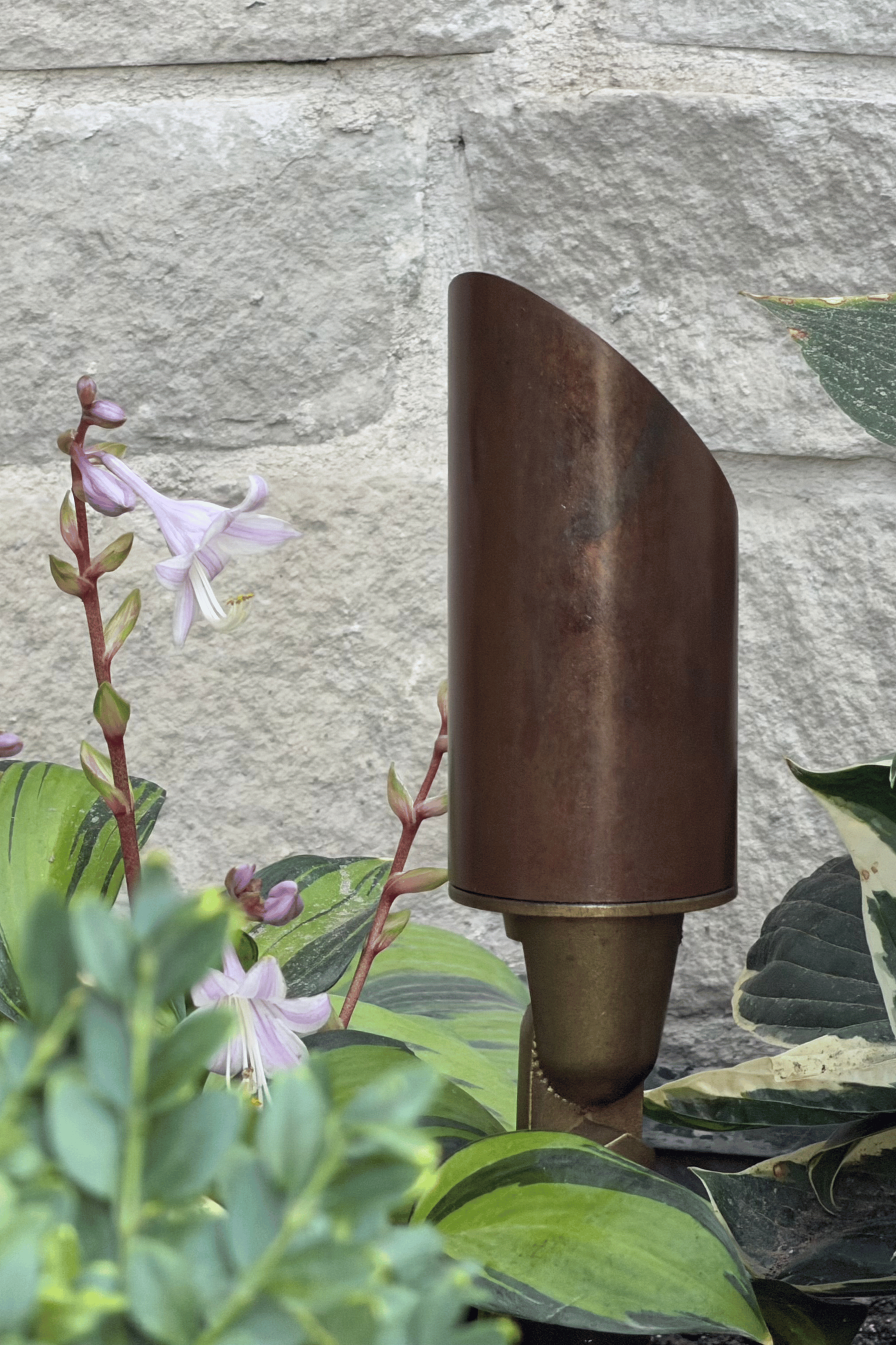 Copper outdoor light mounted on a stone wall, partly framed by pink flowers and green leaves Copper outdoor light mounted on a stone wall, partly framed by pink flowers and green leaves