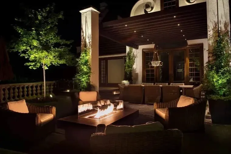 Patio at night with wicker seating around a fire pit, warm lights, and a modern house backdrop