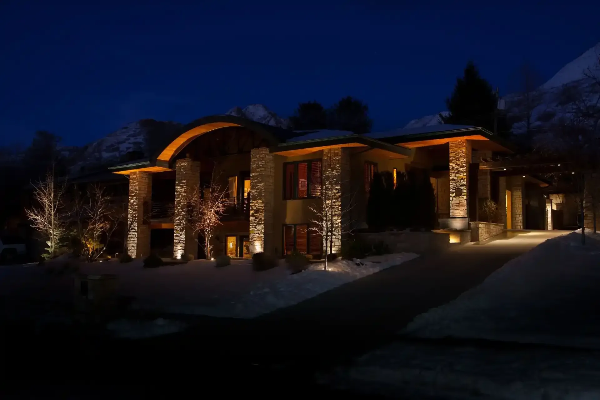 Snowy house at night with warm porch lights and lit stone exterior
