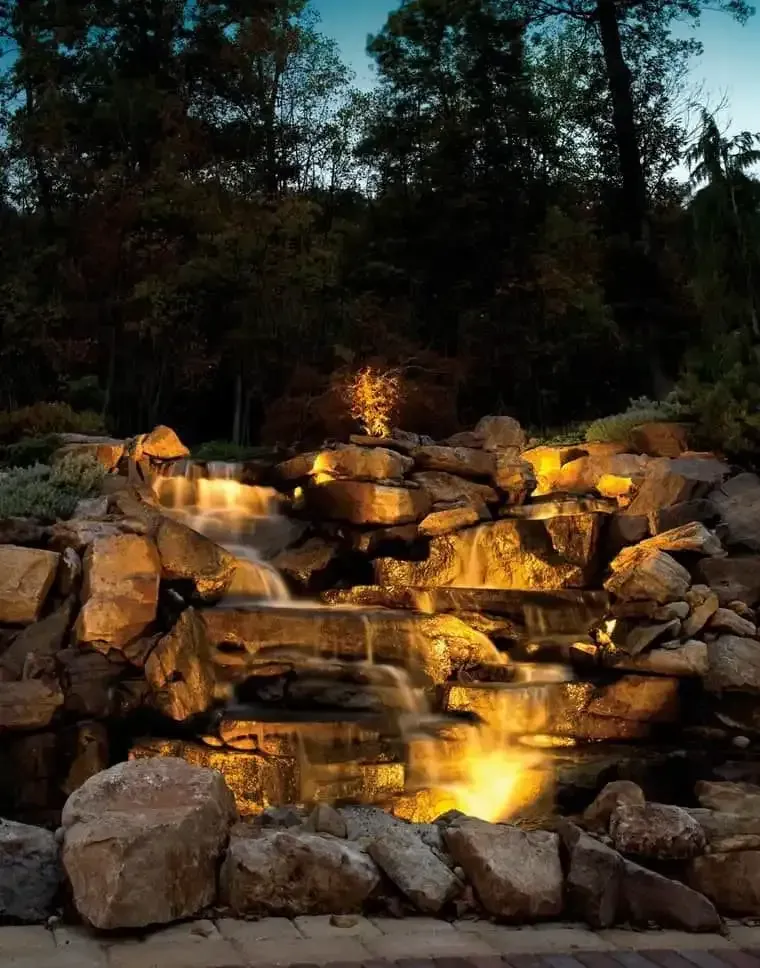 Illuminated backyard waterfall cascading over stacked rocks at dusk, framed by dark trees