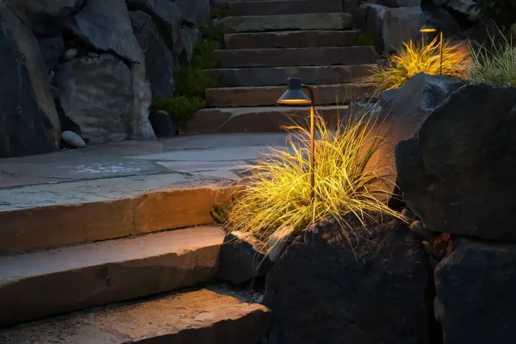 Stone steps at dusk beside rocks, with warm lights illuminating dry ornamental grass.