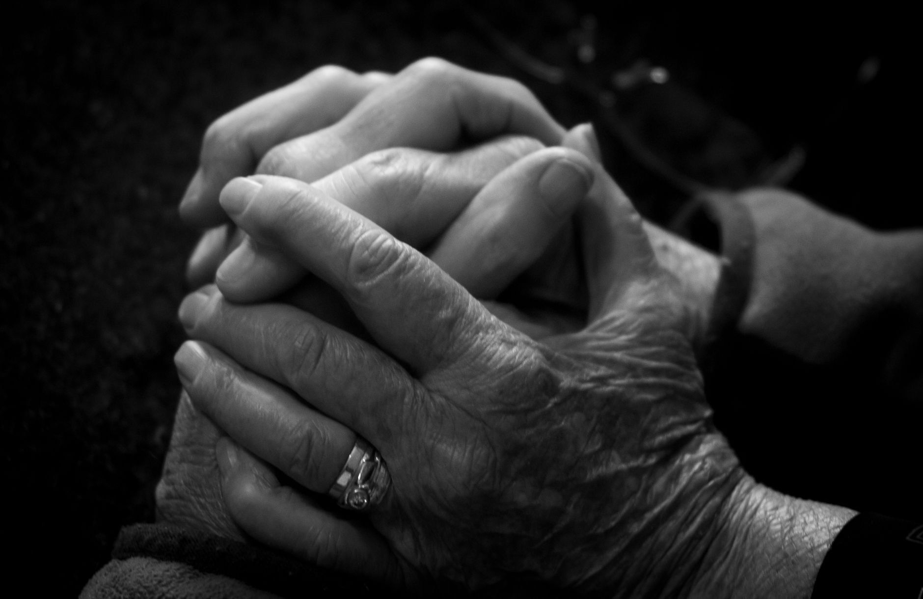 A close-up black-and-white photograph of an elderly person's weathered hands clasped together, with a younger hand resting gently on top, set against a dark, unlit background.