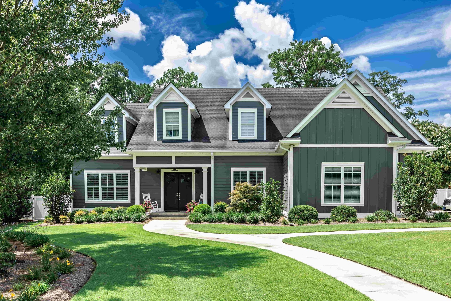 Green house with white trim, dormer windows, and a curved walkway in a sunny landscaped yard.