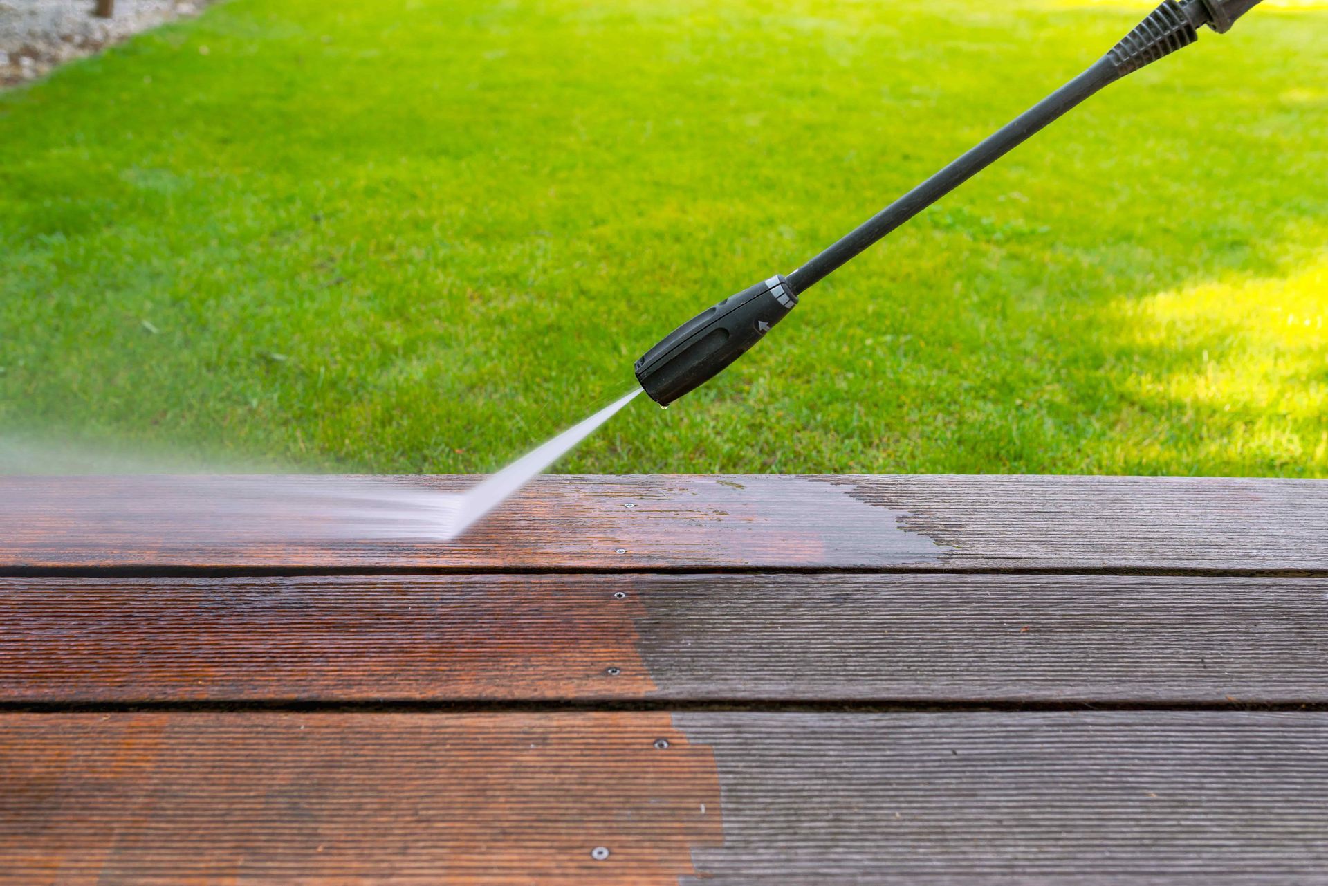 Pressure washing a wooden deck, spraying water across wet brown boards and green grass background