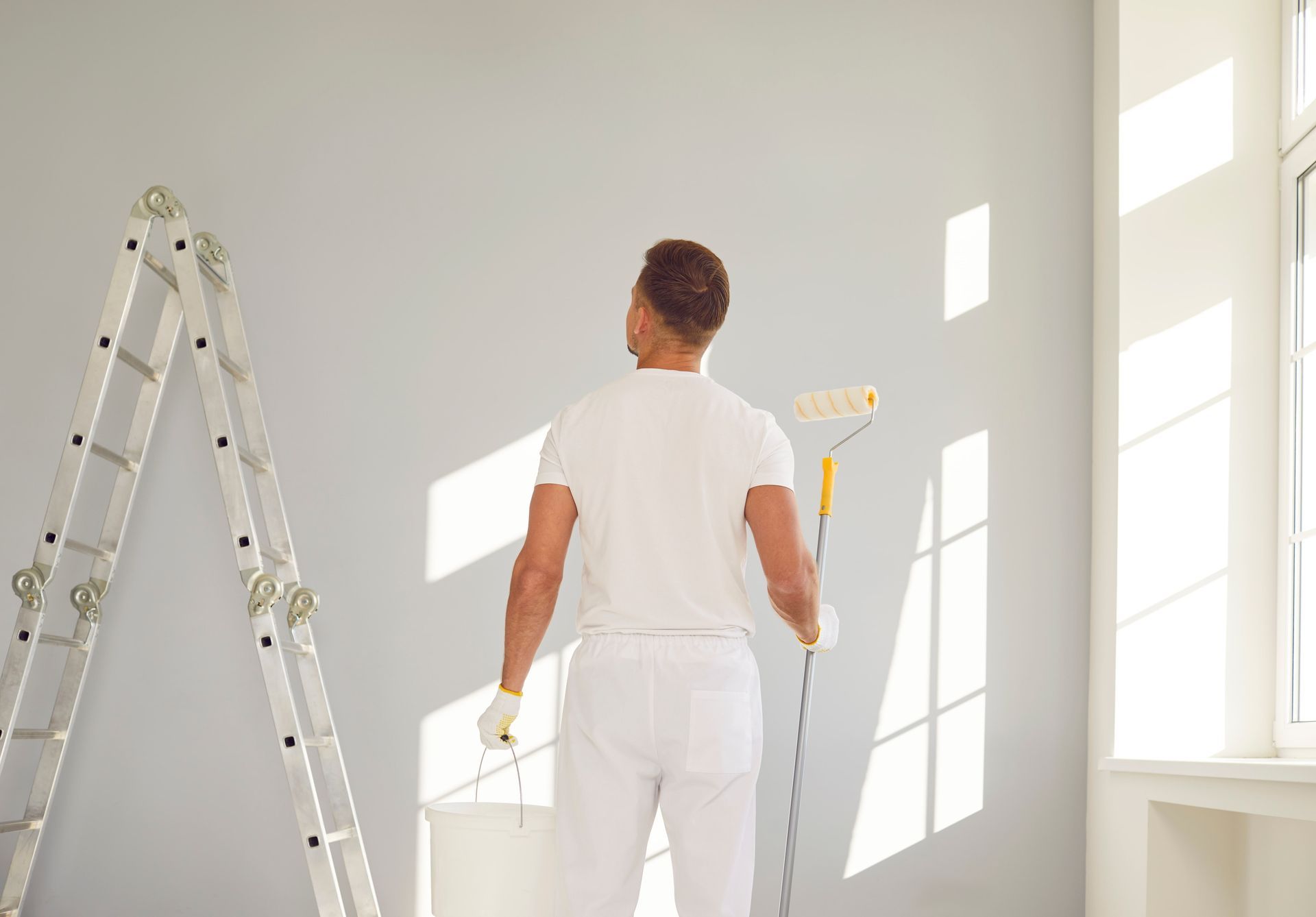Person painting a white wall in a bright room beside a ladder and windows