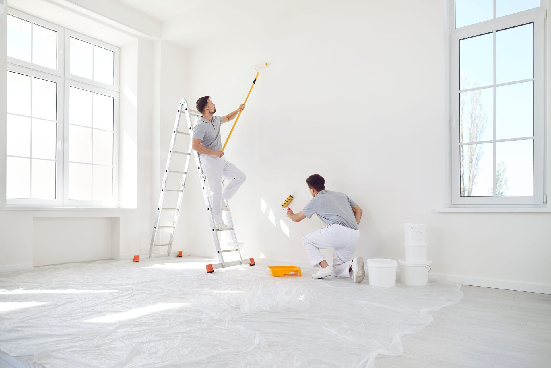 Two people painting a bright white room, one on a ladder and one kneeling with paint supplies.