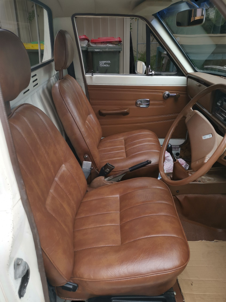 Brown leather car seats inside a vintage vehicle with a wooden door panel and tan steering wheel.