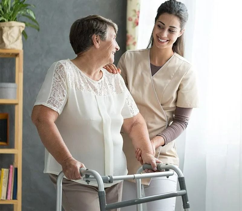 Woman With a Walker Assisted by a Smiling Caregiver Indoors — Ballina Home Healthcare Sales & Hire and Compression Stockings Plus in Ballina, NSW