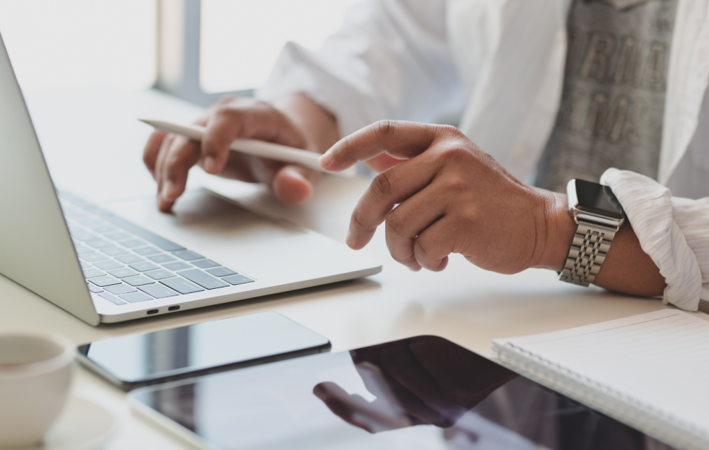 Person using a laptop, tablet, and smartphone at a desk, likely working. Includes pen, watch, and coffee cup.