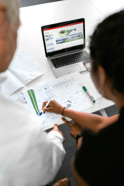 Two people collaborating over architectural plans, laptop on desk, using a blue marker.