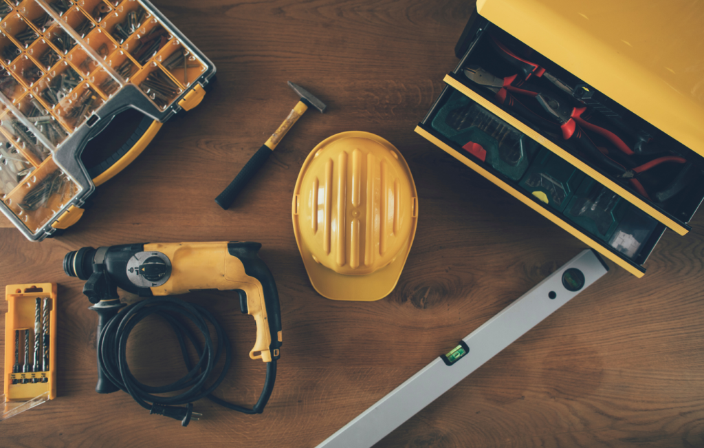 Yellow construction tools: drill, hard hat, level, hammer, toolbox, screw set on wood.
