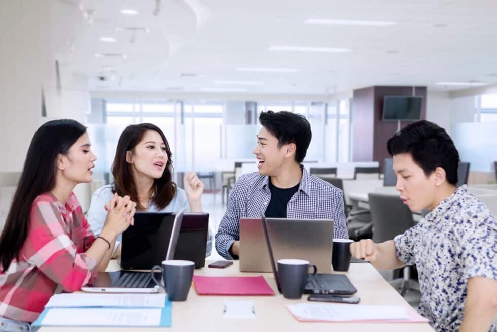 Four diverse young adults at a table with laptops, engaged in animated discussion, in a bright office space.