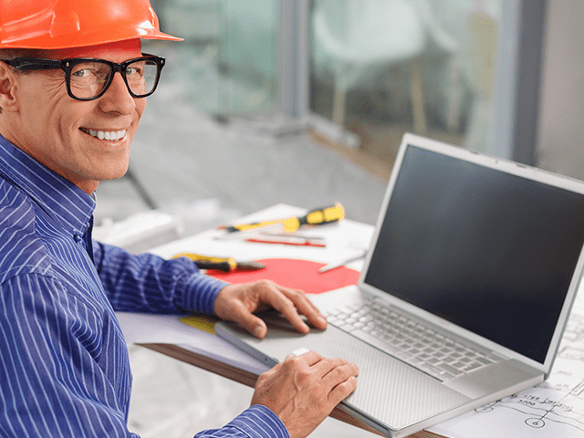 Architect in orange hardhat, smiles while working on laptop with blueprints.