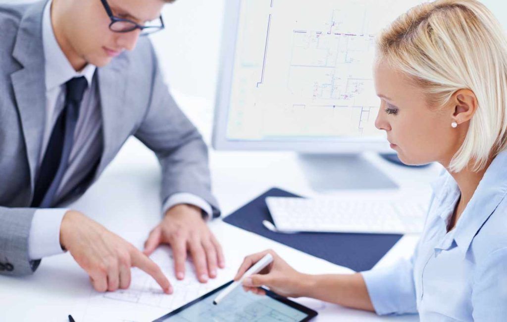 Two business colleagues reviewing documents together at a desk. The man points, woman uses a stylus on a tablet.