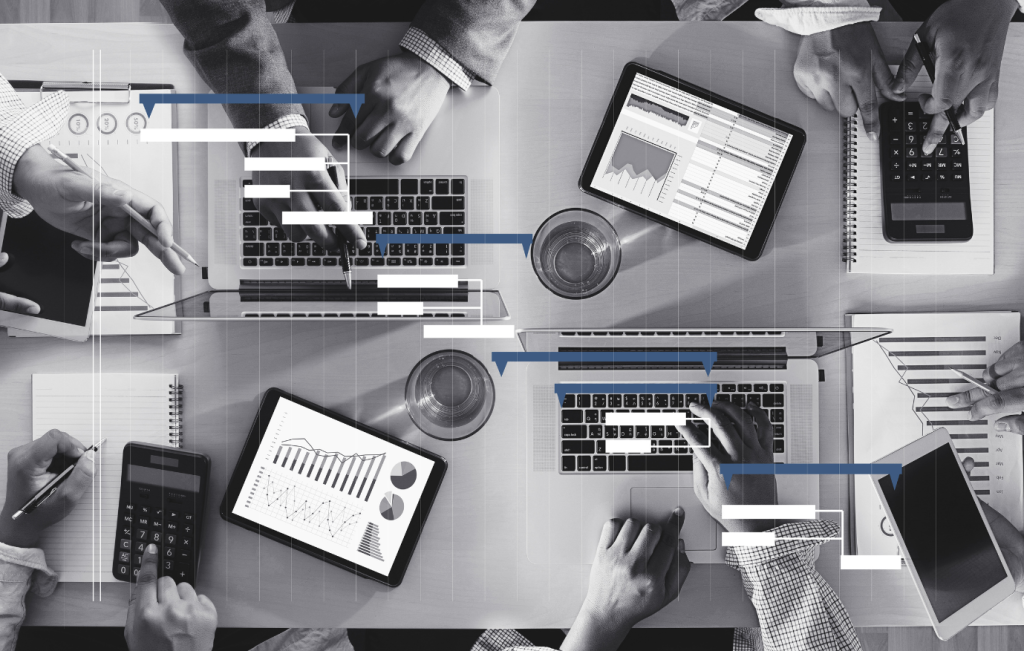 Team working at a table with laptops, tablets, calculators, and papers. Blue lines connect devices, suggesting data sharing.