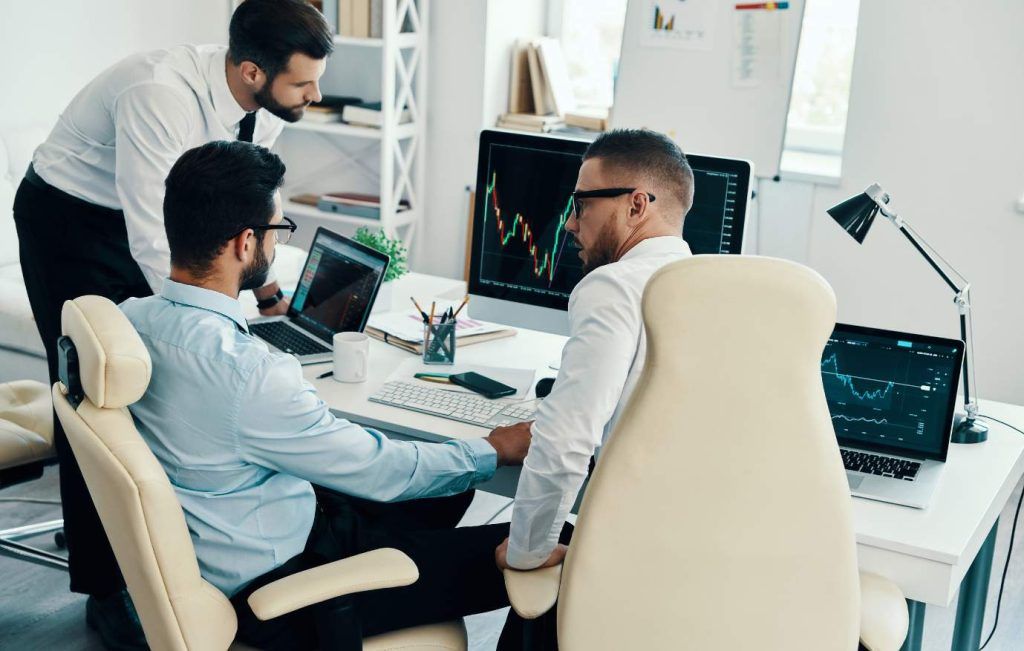 Three businessmen in an office looking at computer screens with financial charts.