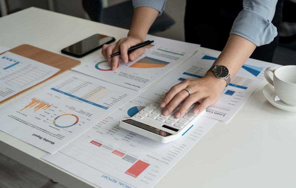 Person analyzing charts with a calculator on a white desk. Includes phone, pen, and a mug.