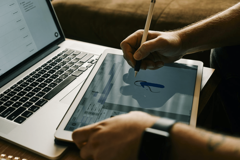 Person using a stylus to draw on a tablet next to a laptop, indoors.