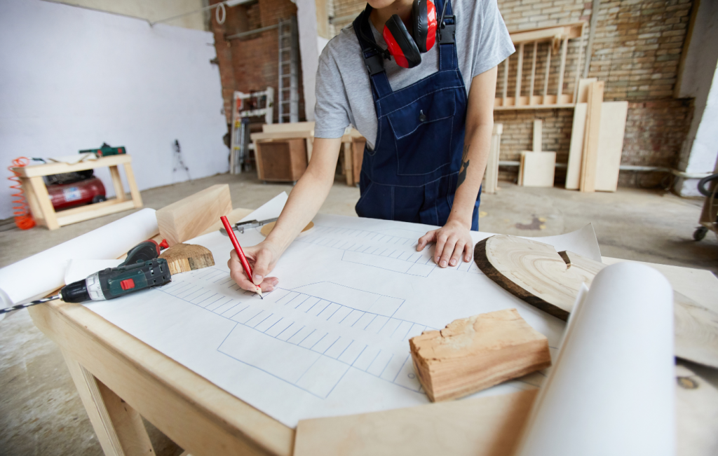Person in overalls sketching blueprints on a workbench in a woodworking shop.