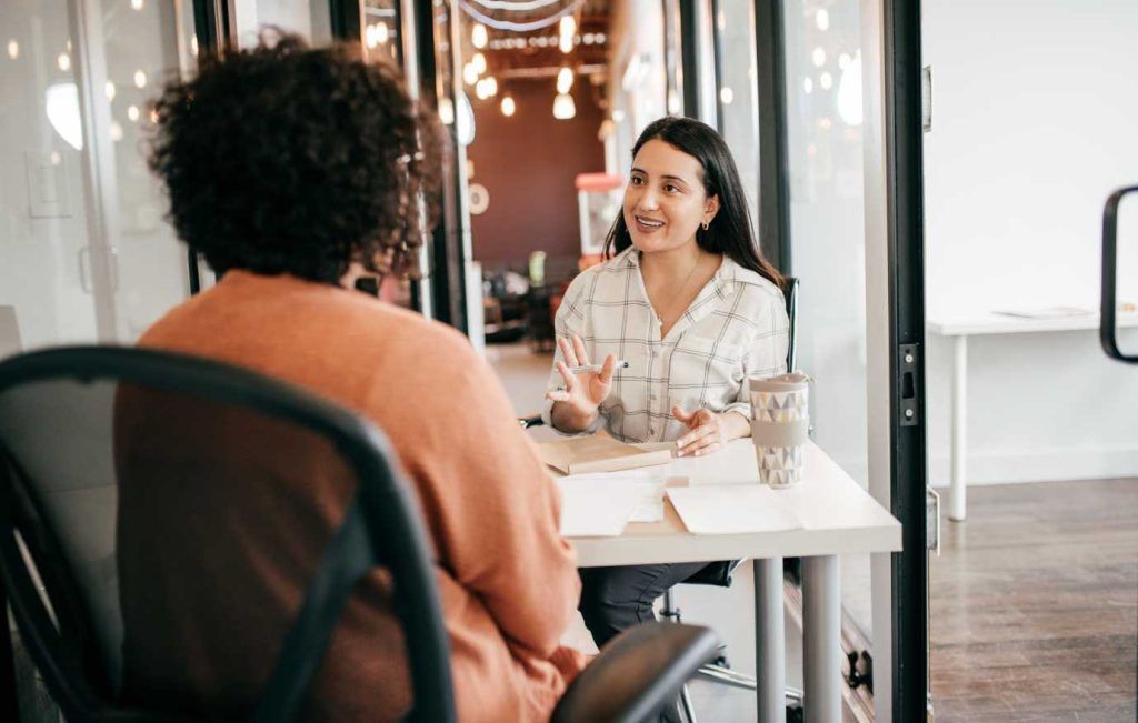 Two women at a table, one speaking, the other listening; they are in an office setting.