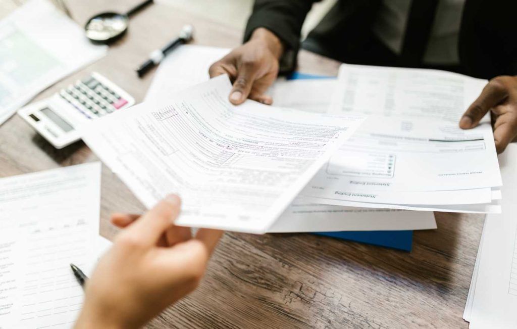 Hands exchanging documents at a desk, a calculator and magnifying glass visible.