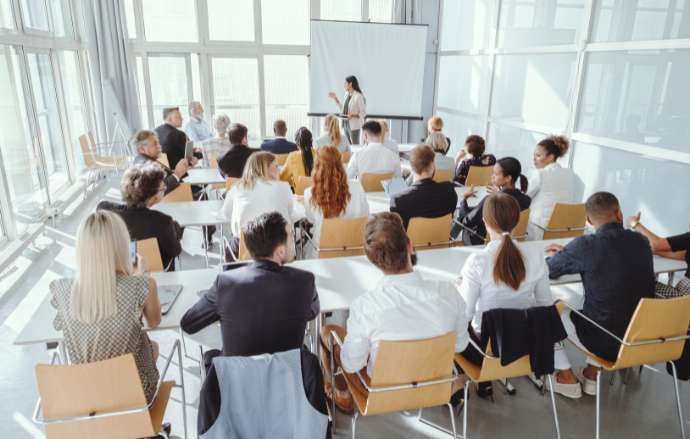 A woman presents to a diverse group in a bright conference room; attendees sit at tables, looking toward the screen.