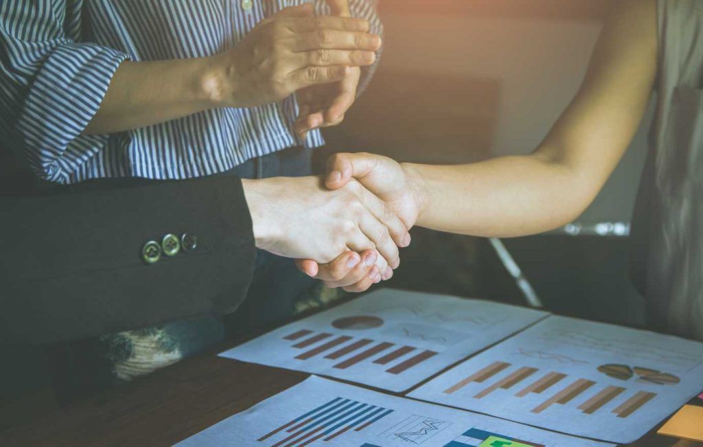 Two people shaking hands over documents with graphs.