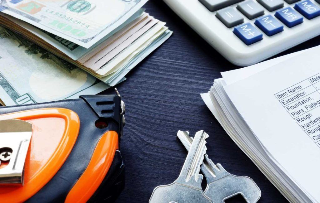 Stack of cash, keys, calculator, tape measure, and papers on dark surface.