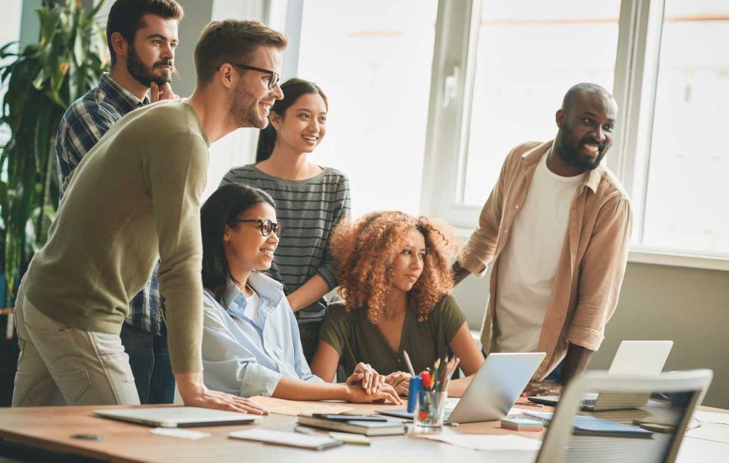 Group of diverse coworkers gathered around a laptop in a bright office, smiling and collaborating.
