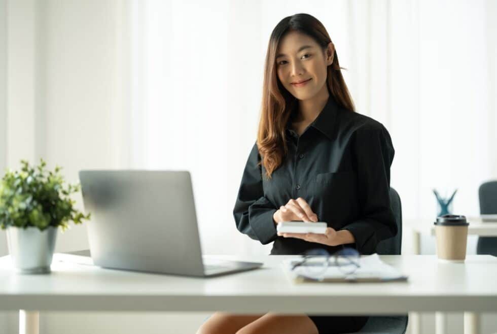 Woman in black shirt at desk, using calculator, smiling at camera. Office setting.