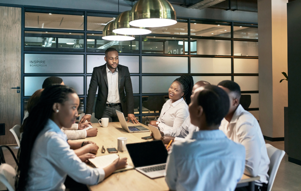 Group of diverse professionals in a meeting; a man stands at the head of the table, addressing the group.