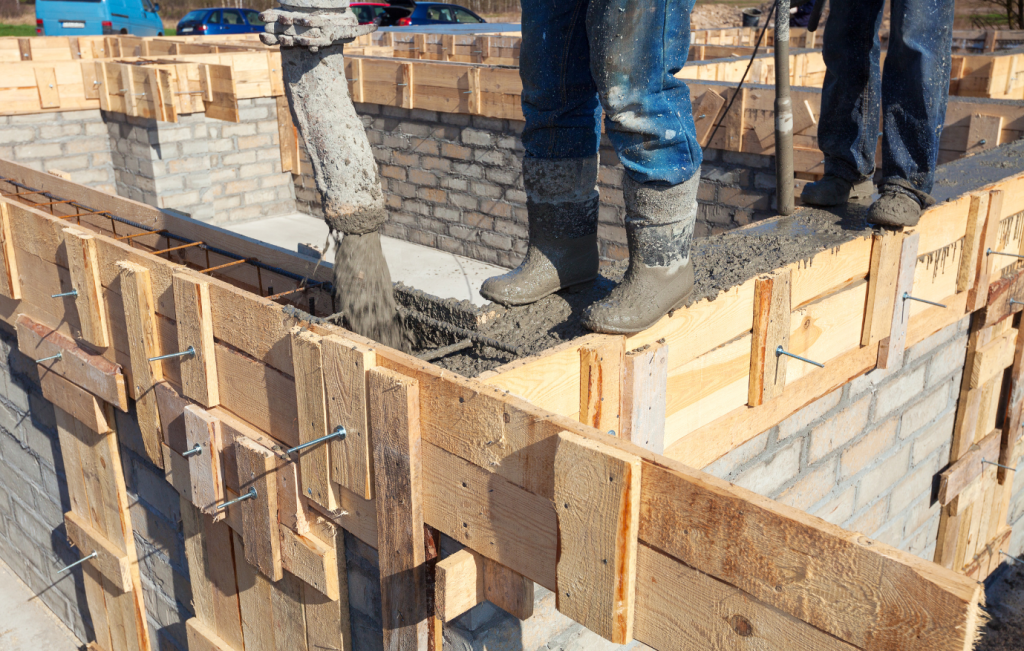 Construction workers pouring concrete into wooden forms on a brick foundation.