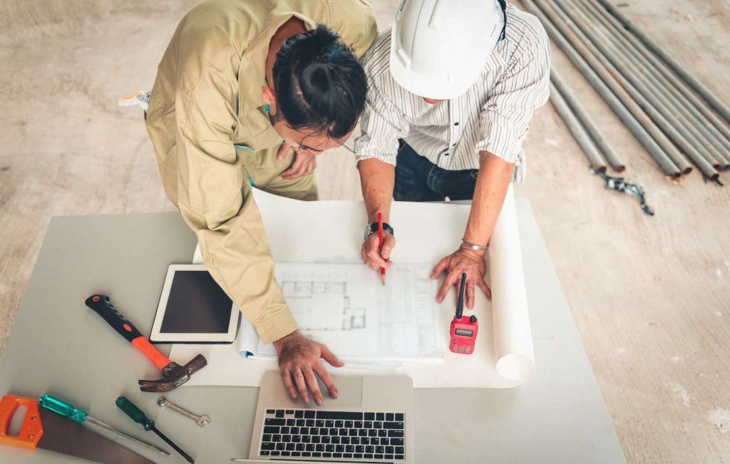 Two construction workers reviewing blueprints at a table with tools.