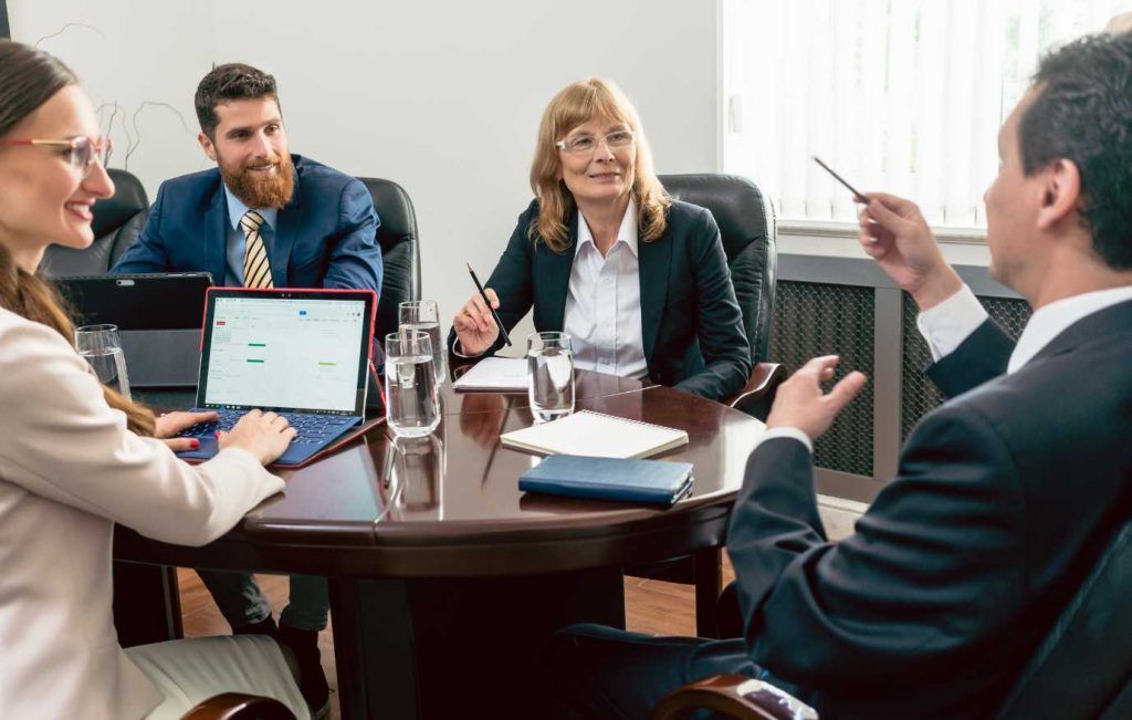 Four business people in a conference room, one points upward, others look on, smiling.