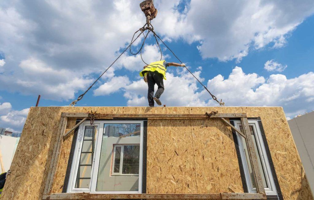 Construction worker guiding a modular wall section with windows being lifted by a crane against a blue sky.