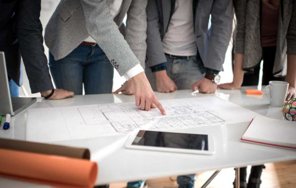 Team members lean over a table, pointing at papers and a tablet; collaborative planning.
