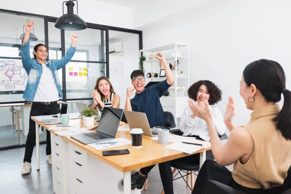 Team celebrates success in bright office. People cheering and clapping around a table with laptops.
