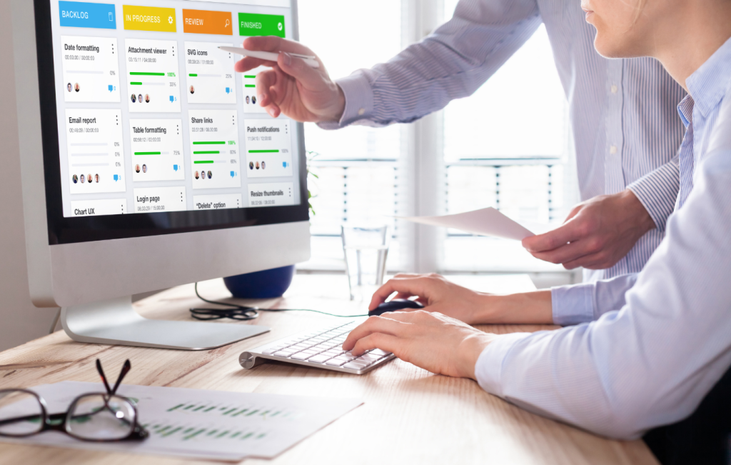 Two people at a desk reviewing project management software on a computer screen.