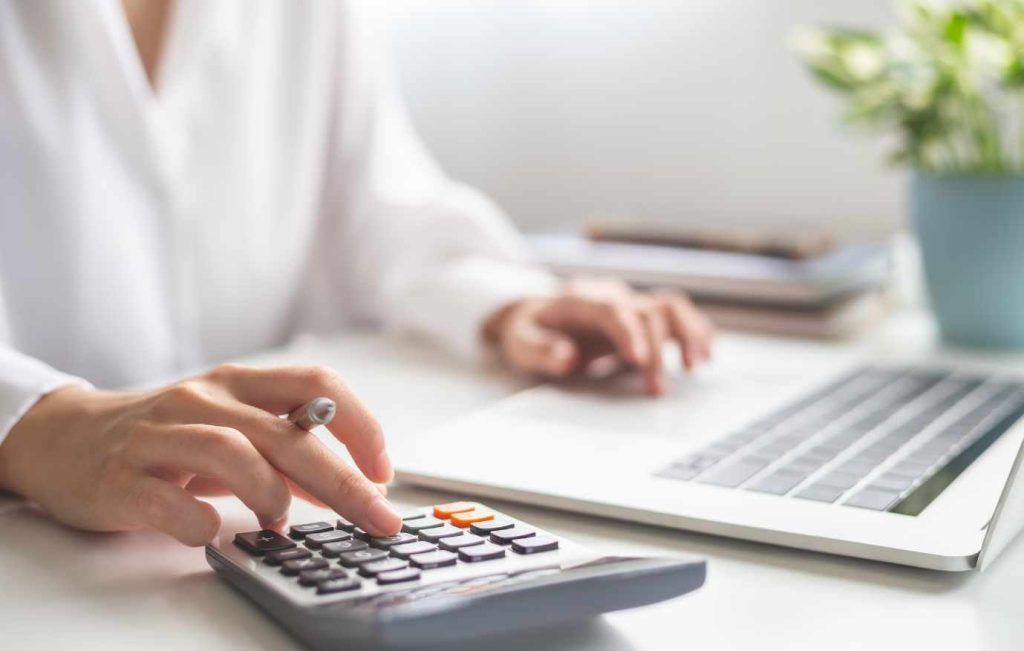 Woman's hands using a calculator on a white desk, laptop, and plant in background.