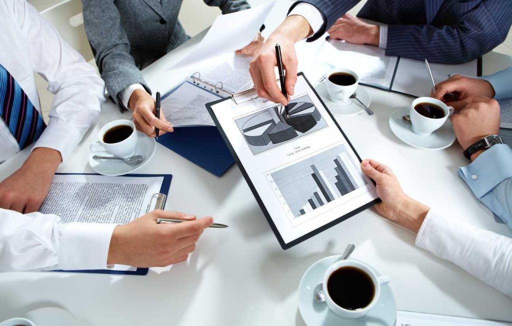 Business meeting around a white table, reviewing graphs and documents with coffee.