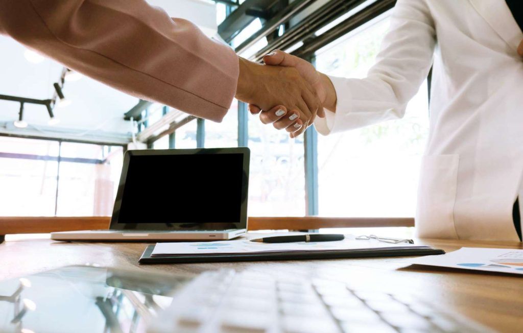 Two people shaking hands over a desk with a laptop and documents.