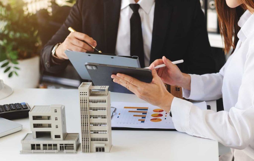 Two people in suits reviewing documents over a model of buildings, possibly a real estate or investment meeting.