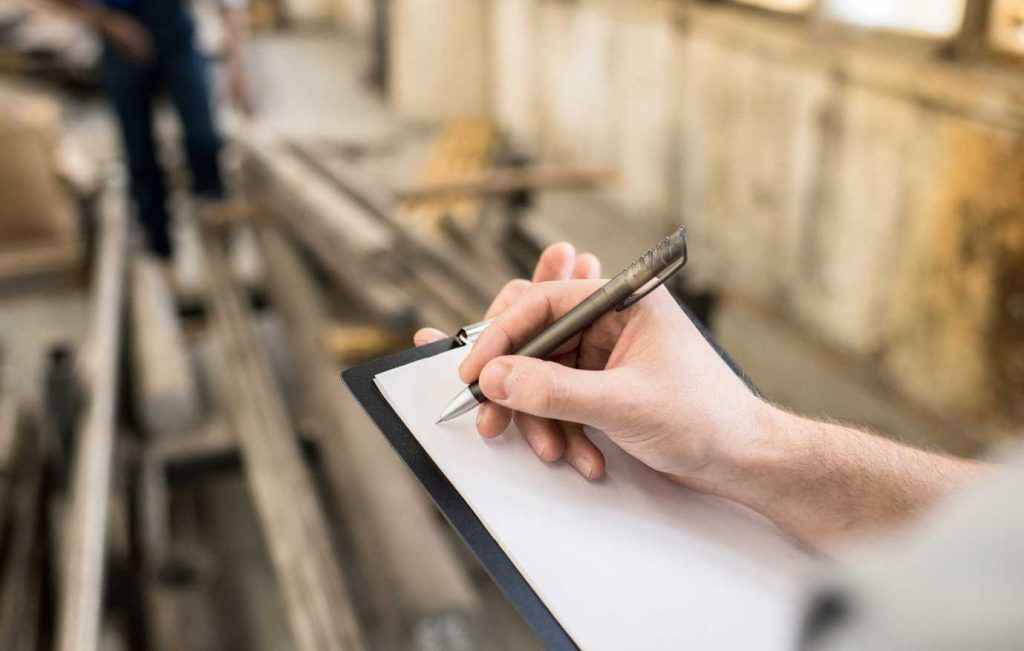 Person writing on a clipboard in a wood shop, surrounded by lumber.
