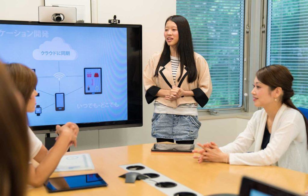 Woman giving a presentation in a meeting room, standing by a large screen. Others listen at the table.