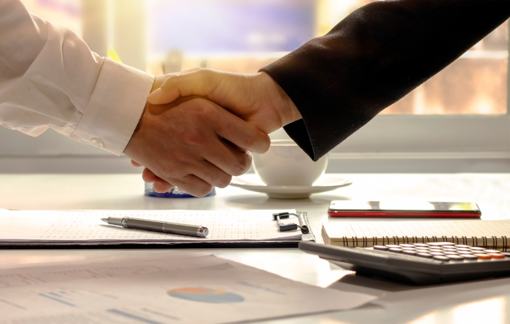Business handshake over a desk, finalizing a deal. Papers, pen, calculator, and coffee cup present.