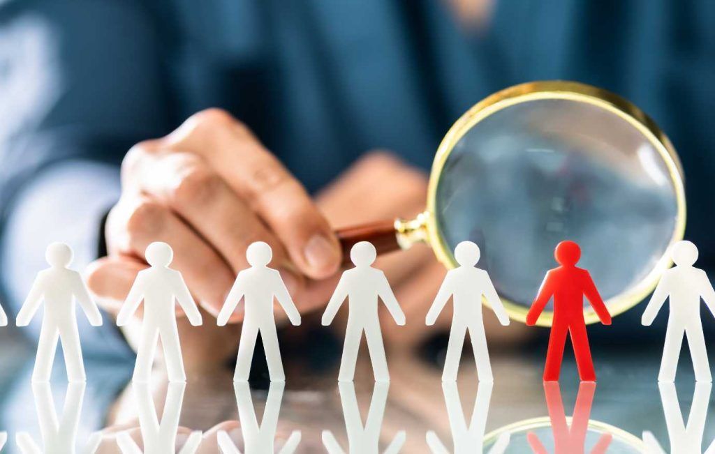 Hand holding magnifying glass over a red figure amidst white figures; recruitment concept.
