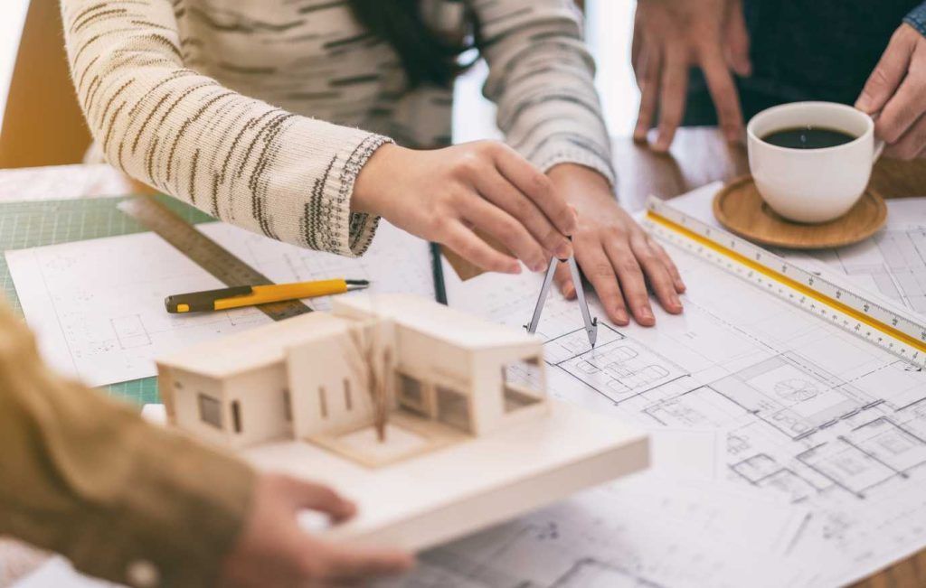 Architects collaborating at a table; one uses a compass on blueprints, a model house and coffee cup present.